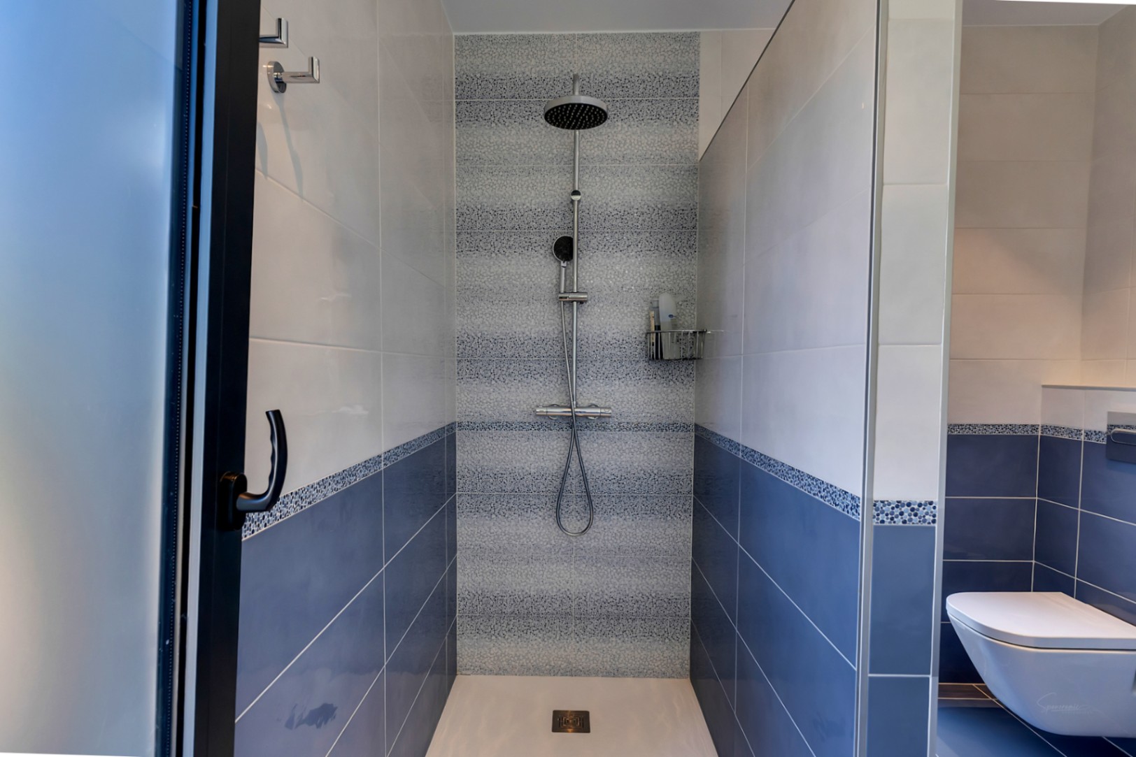 Ducha de obra con azulejos bicolores en azul y blanco, y una franja decorativa de mosaico. Equipada con rociador de lluvia y ducha de mano, separada por mamparas de cristal.
