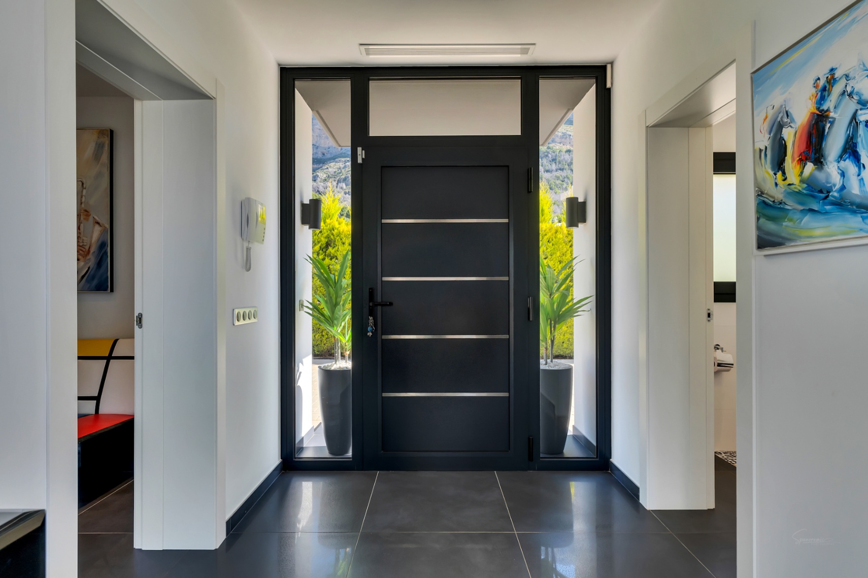 Elegante recibidor con moderna puerta oscura, paneles de cristal laterales y superior, que ofrece luz natural y vistas al exterior y montañas. Suelo de baldosas oscuras e intercomunicador.