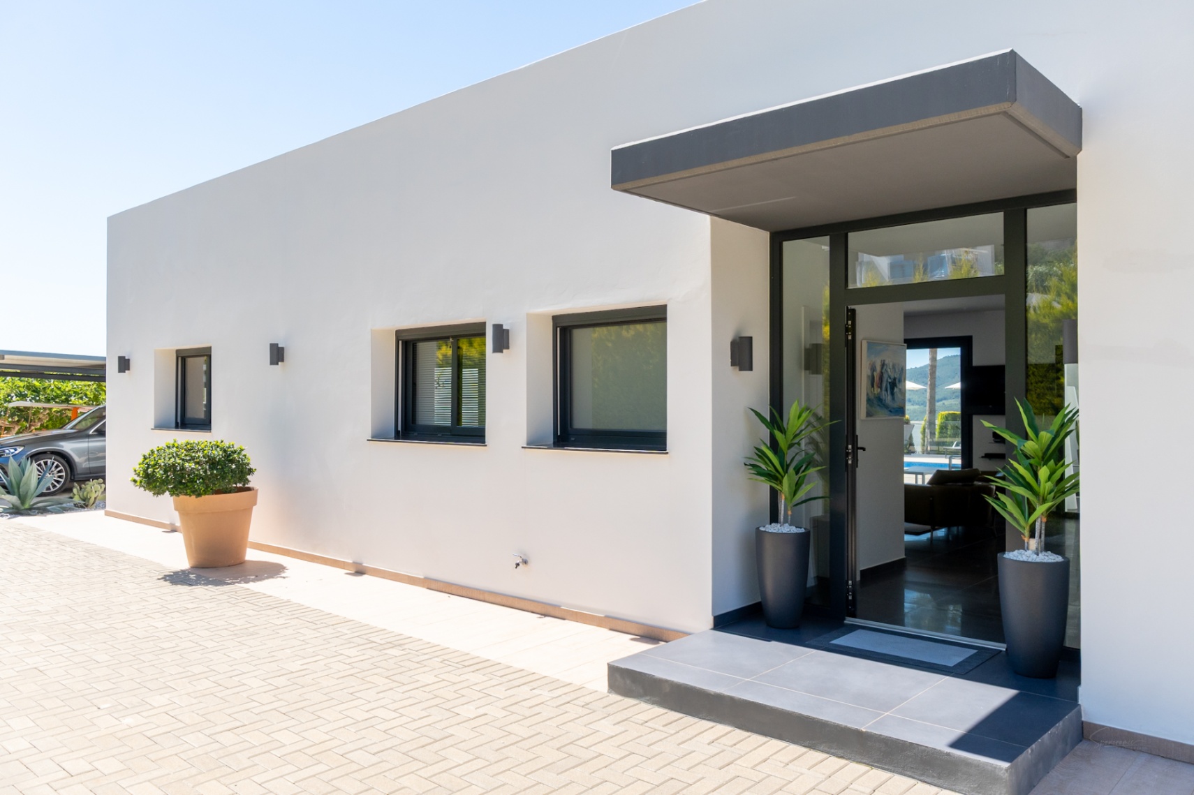 Entrada principal de villa moderna con fachada blanca, puerta acristalada y toldo oscuro. Adoquinado exterior, ventanas con persianas y aparcamiento cubierto visible.