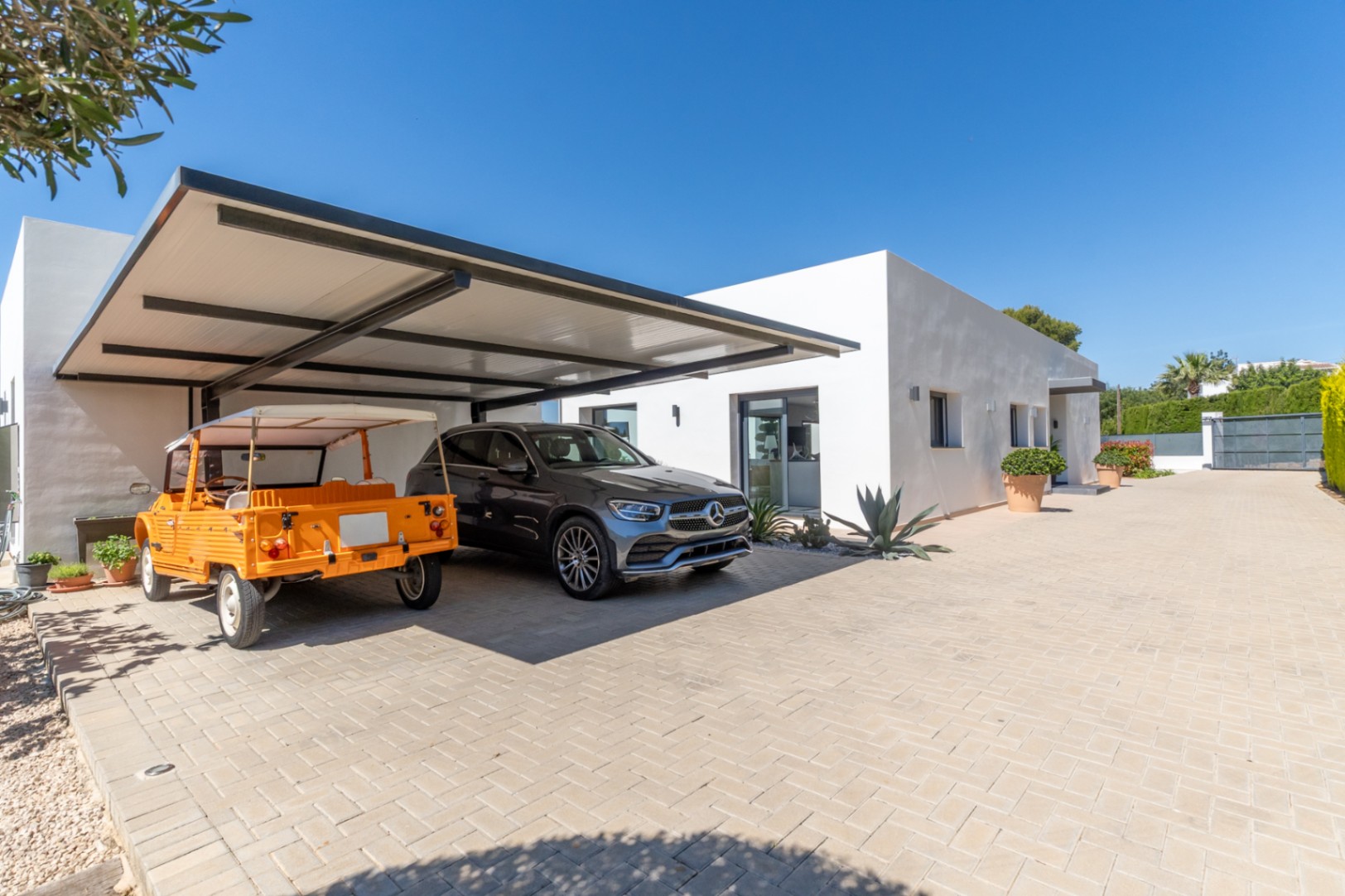 Exterior de villa moderna con doble aparcamiento cubierto de estructura oscura, albergando un SUV Mercedes y un coche descapotable naranja, sobre un camino pavimentado.