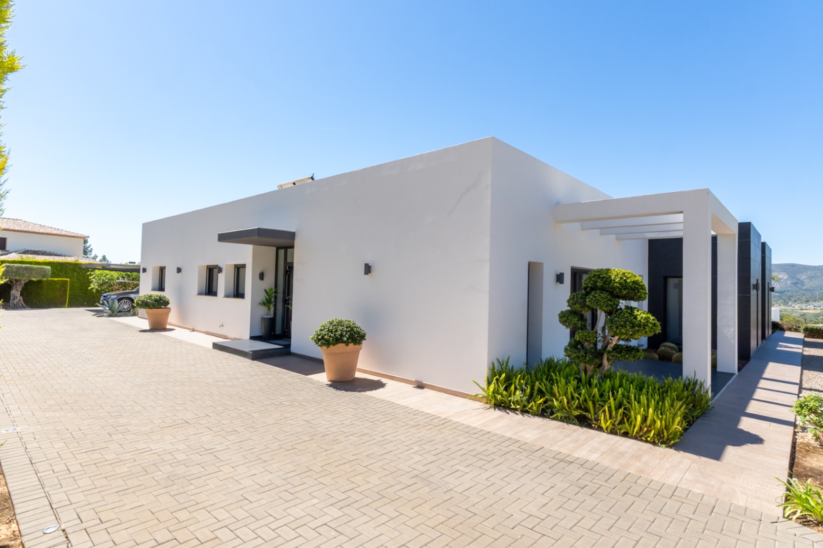 Exterior de villa moderna con fachada blanca, entrada pavimentada y paisajismo minimalista. Destacan un árbol esculpido, pérgola y vistas a las colinas.