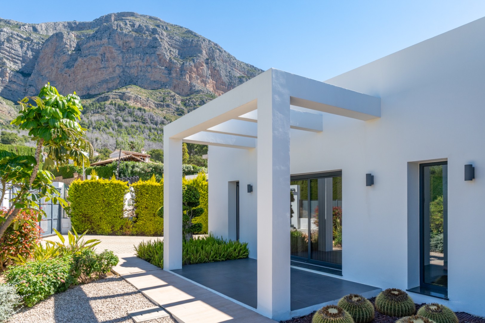 Exterior moderno con pérgola blanca, jardín mediterráneo y vistas despejadas a la majestuosa montaña del Montgó bajo un cielo azul.
