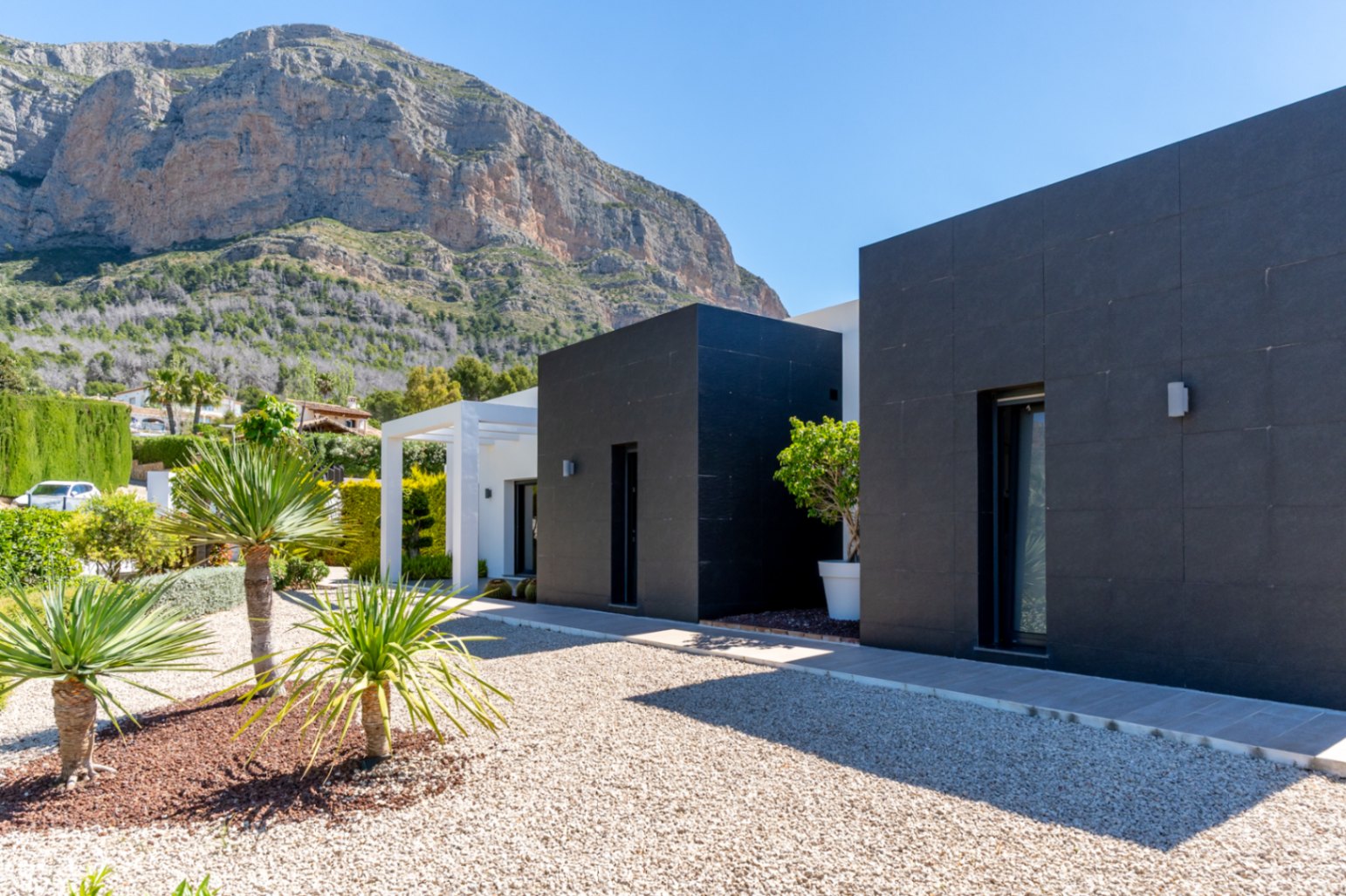 Exterior moderno de la villa con fachadas de azulejos oscuros y detalles en blanco, enmarcada por el imponente Montgó. Jardín de grava con plantas mediterráneas.