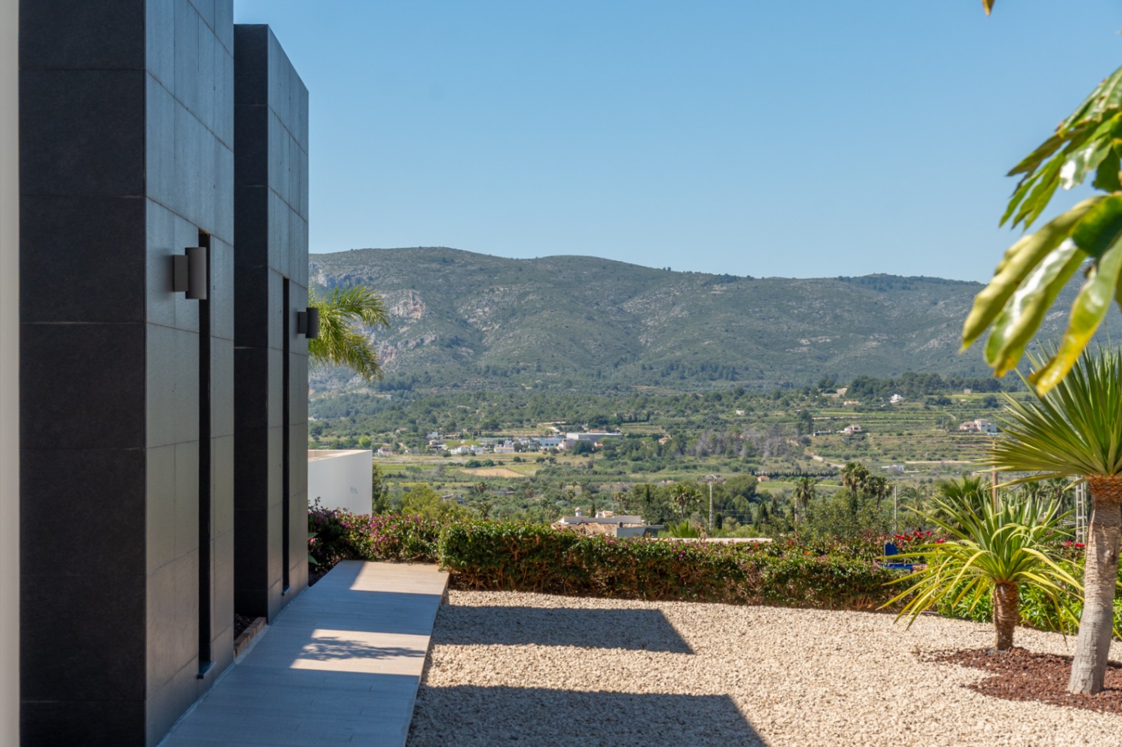 Fachada exterior con revestimiento de paneles oscuros y apliques de pared, sendero pavimentado, jardín de grava y amplias vistas a las montañas del Montgó.
