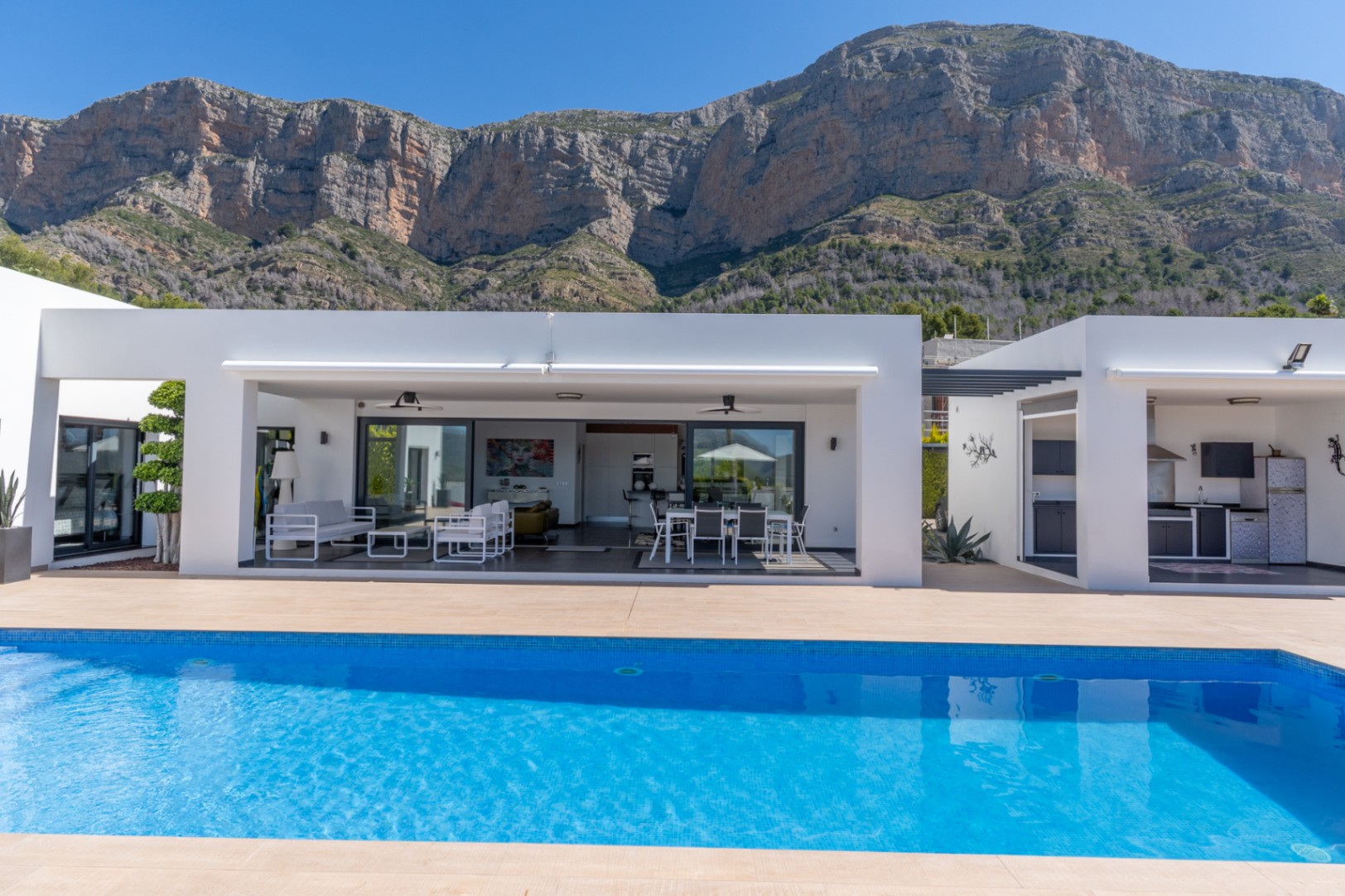 La moderna villa presenta una piscina azul brillante, terraza cubierta con salón y comedor exterior, además de cocina integrada al aire libre, con vistas al Montgó.