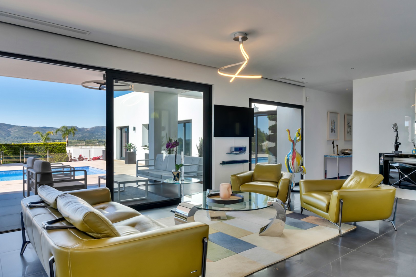 Luminoso salón con sofá y sillones de cuero verde oliva, mesa de centro de cristal y acceso directo a la terraza, piscina y vistas a la montaña.