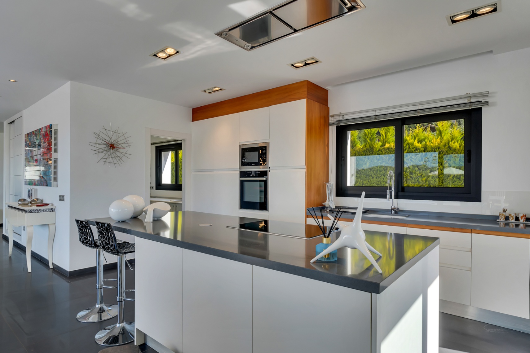 Moderna cocina con gran isla central de encimera oscura y placa integrada, armarios blancos sin tiradores y electrodomésticos empotrados. Ventana amplia con vistas a la vegetación.