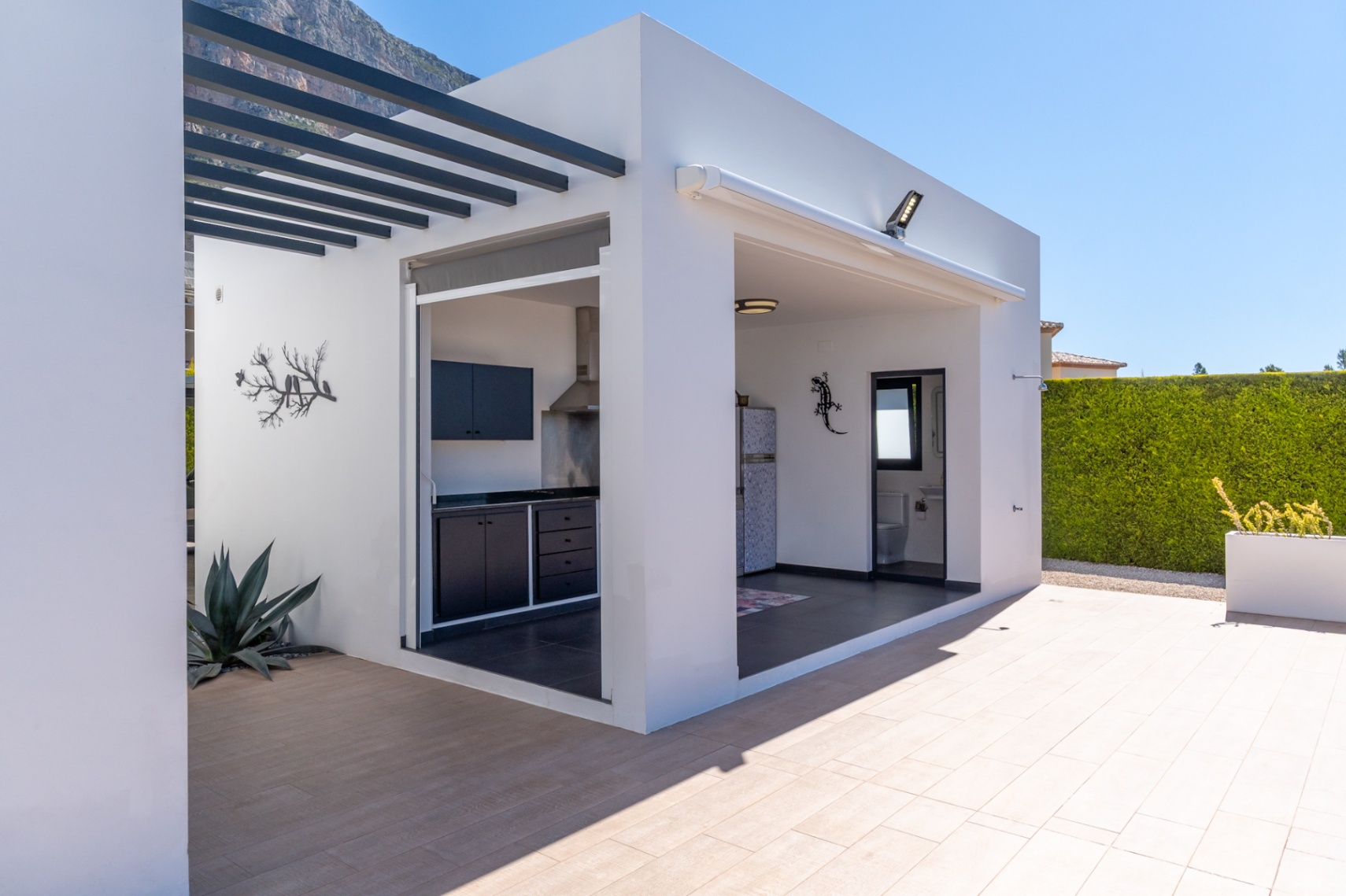 Moderna cocina exterior y zona de barbacoa con mobiliario oscuro, integrada con un práctico baño exterior, todo en un patio embaldosado con vistas a la montaña.