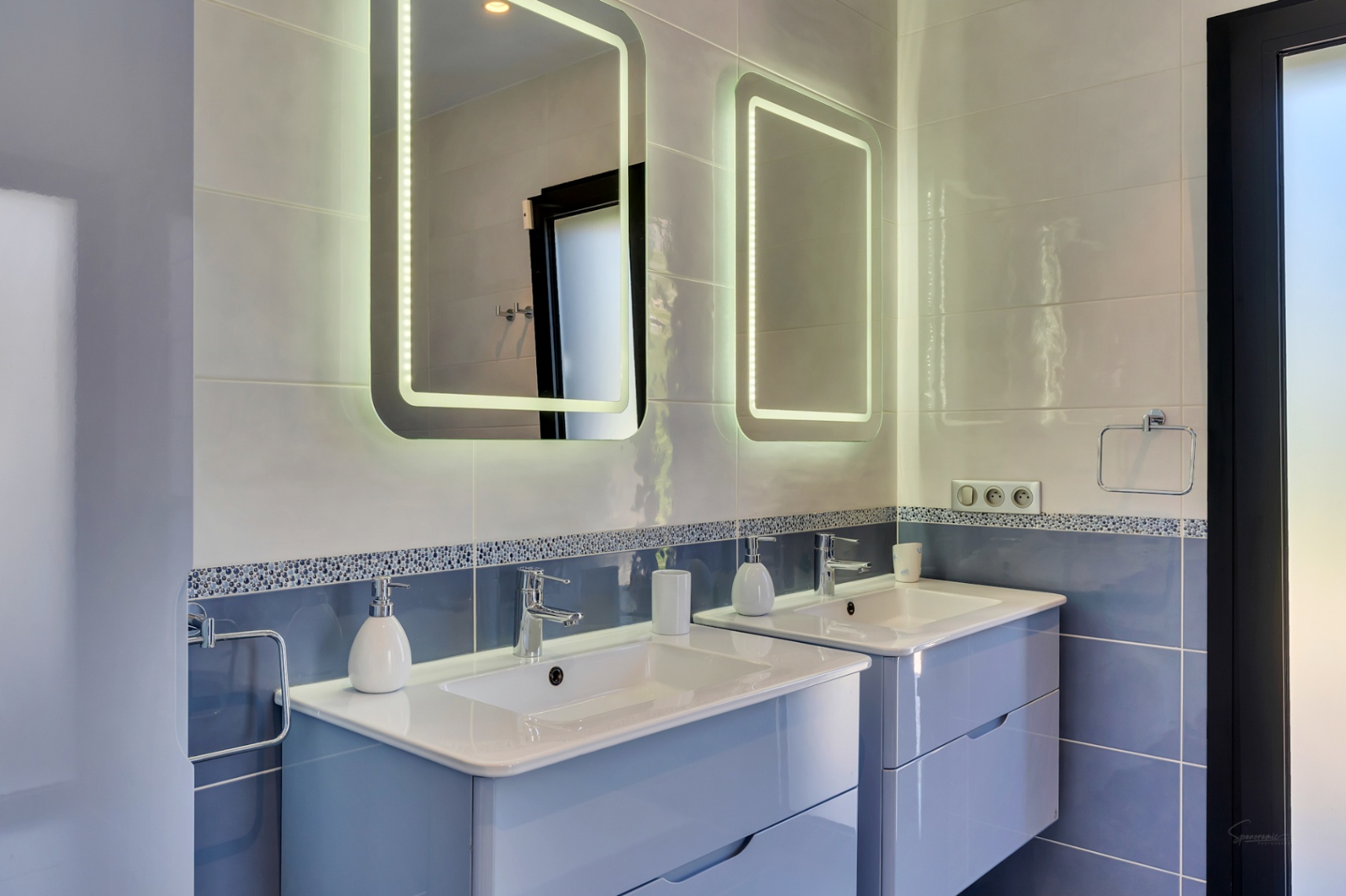 Moderno baño con doble lavabo suspendido de color azul claro, encimeras blancas y dos espejos retroiluminados LED sobre paredes alicatadas en tonos azules y grises.