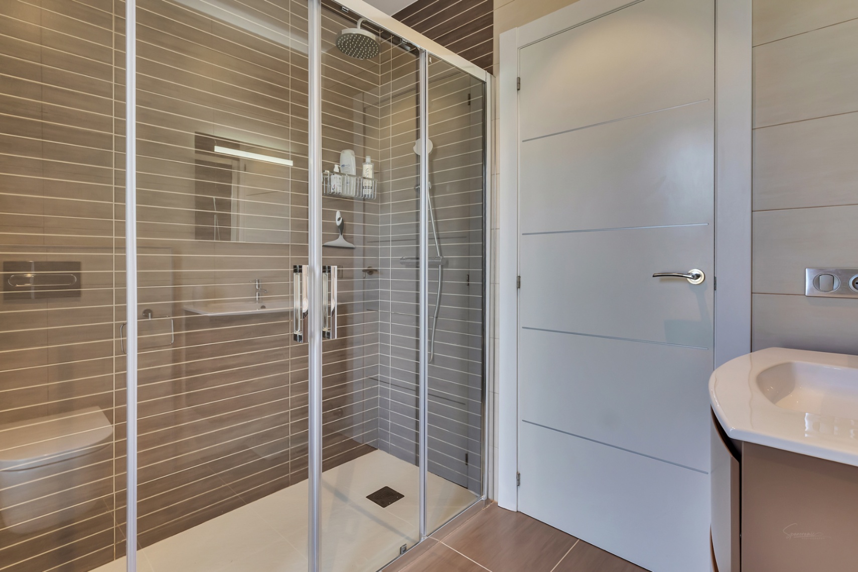 Moderno cuarto de baño con amplia ducha acristalada, cabezal de lluvia y azulejos taupe con rayas horizontales. Incluye inodoro suspendido y lavabo con mueble contemporáneo.