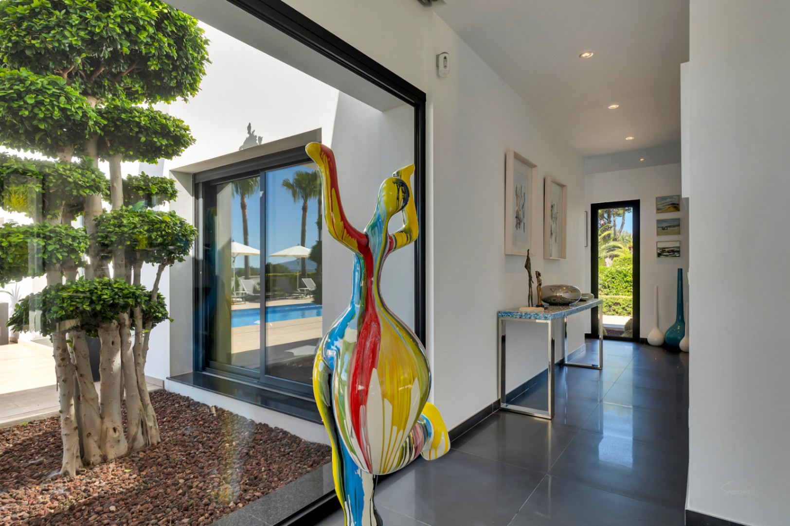 Moderno pasillo con suelo oscuro, escultura abstracta colorida y vistas a la terraza con piscina y un árbol ornamental interior.