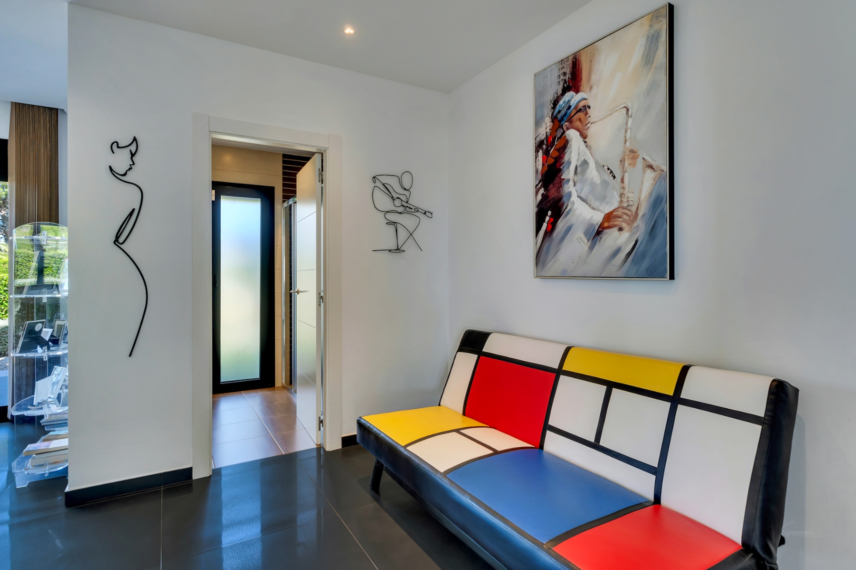 Pasillo moderno con sofá de diseño Mondrian, cuadro de saxofonista y arte de alambre minimalista, sobre suelo de baldosas oscuras y brillantes.