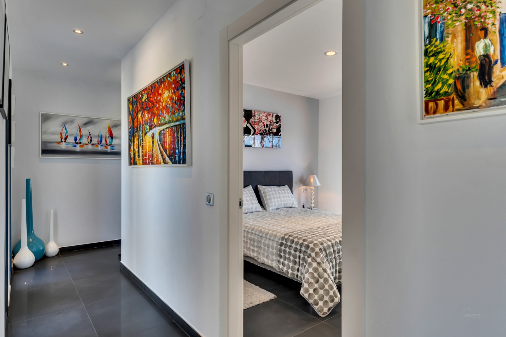 Pasillo moderno con suelo de gres oscuro, iluminación empotrada y vibrante arte abstracto, que conduce a un dormitorio con cama estampada y lámpara de noche.