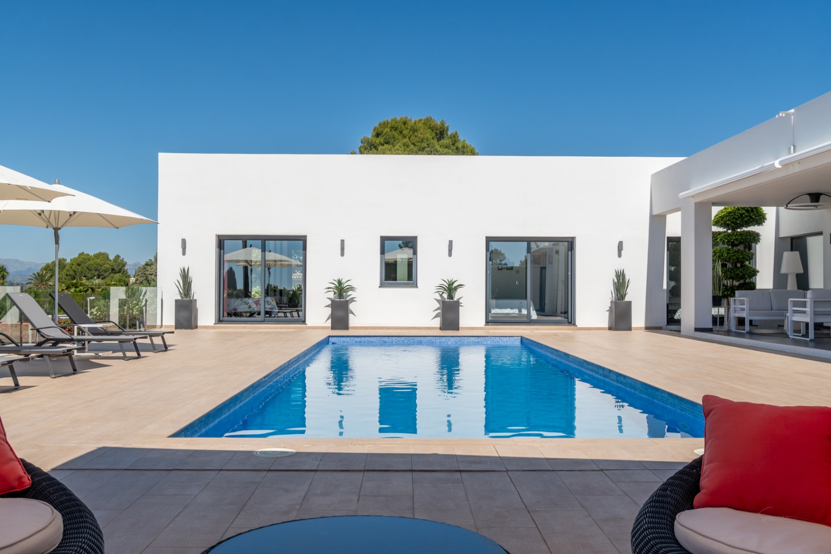 Piscina rectangular de mosaico azul rodeada de una terraza de gres, frente a una moderna fachada blanca con amplias puertas correderas y un salón exterior cubierto.