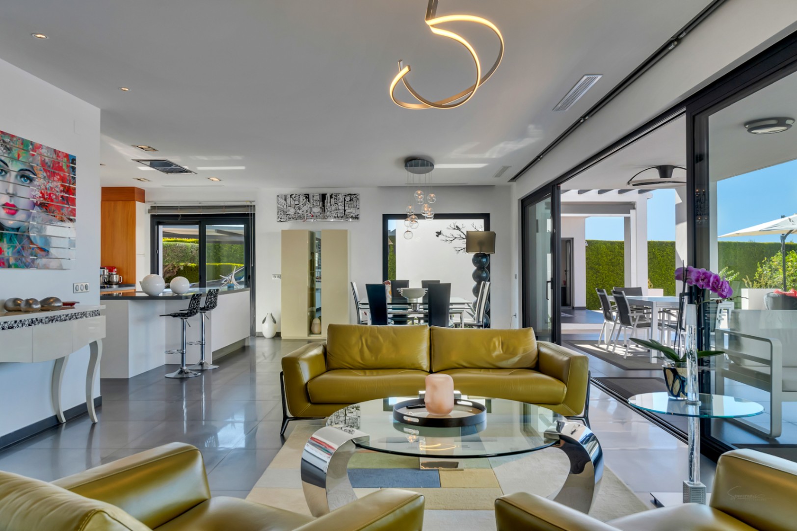 Salón de planta abierta con sofás de piel verde oliva, mesa de centro de cristal y acceso directo a la terraza exterior mediante grandes puertas correderas. Iluminación moderna.
