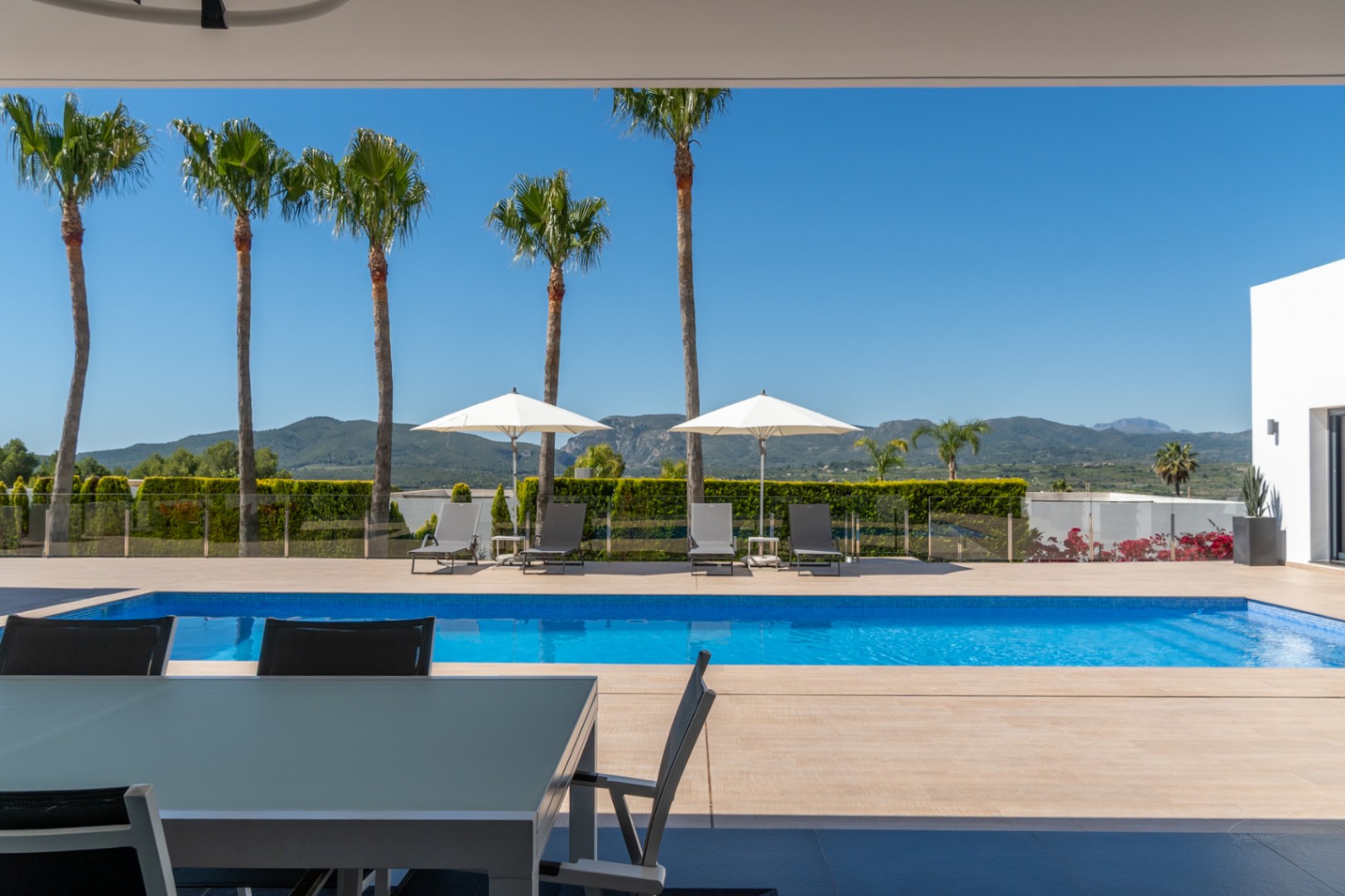 Terraza cubierta con mesa de comedor exterior, con vistas a la piscina de azulejos azules y al paisaje montañoso con palmeras bajo un cielo despejado.