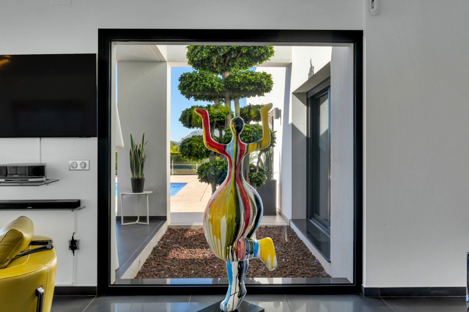 Una escultura moderna y colorida enmarca la vista desde el salón a través de un gran ventanal negro hacia un jardín cuidado con un árbol escalonado y piscina.