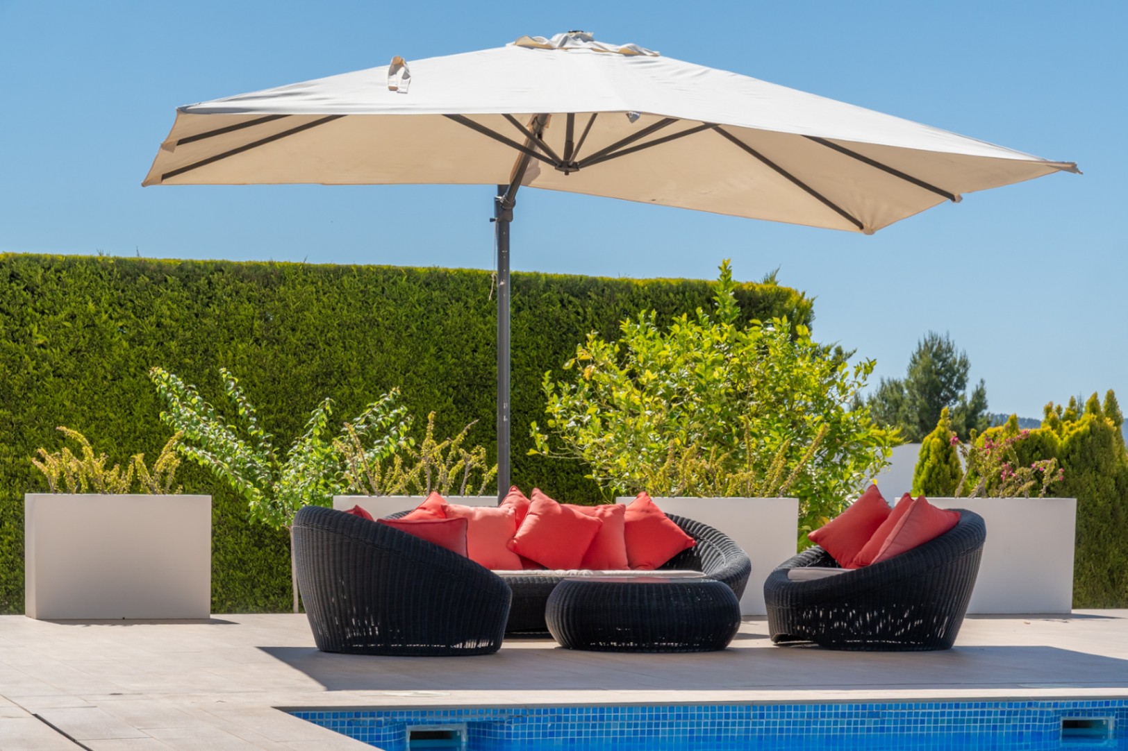 Zona de estar exterior con mobiliario de ratán sintético negro y cojines rojos, bajo una sombrilla, junto a la piscina de mosaico azul.