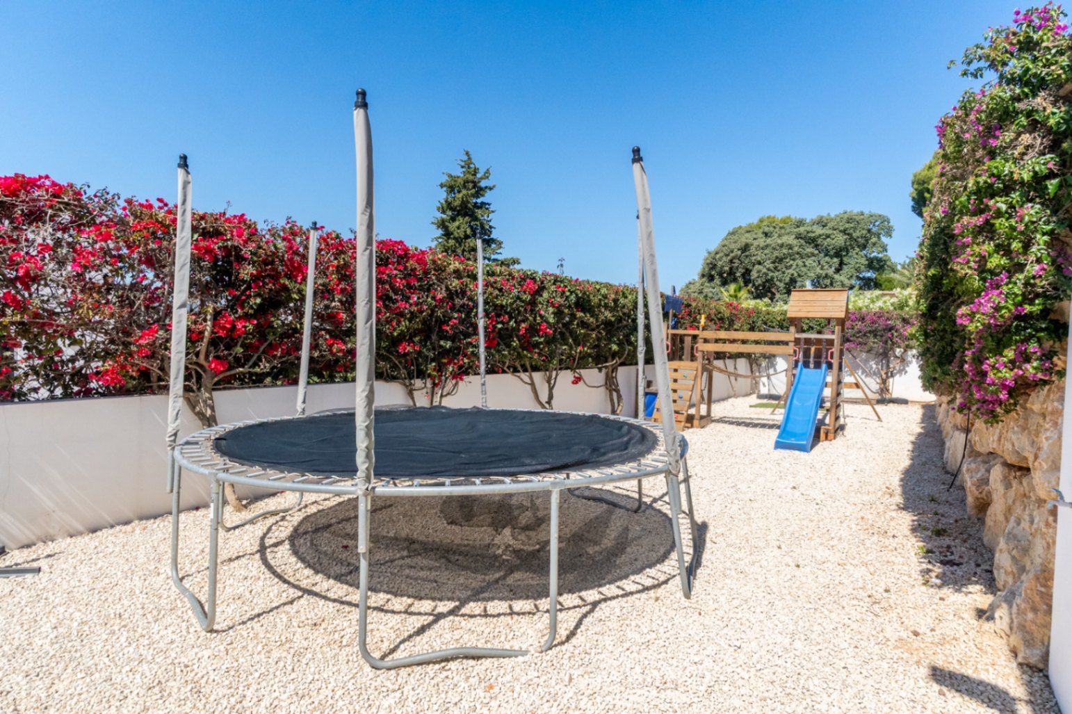 Zona de juegos exterior con cama elástica y estructura de madera con tobogán azul sobre gravilla. Arbustos de buganvilla y un muro de piedra con enredaderas moradas enmarcan el espacio bajo un cielo despejado.