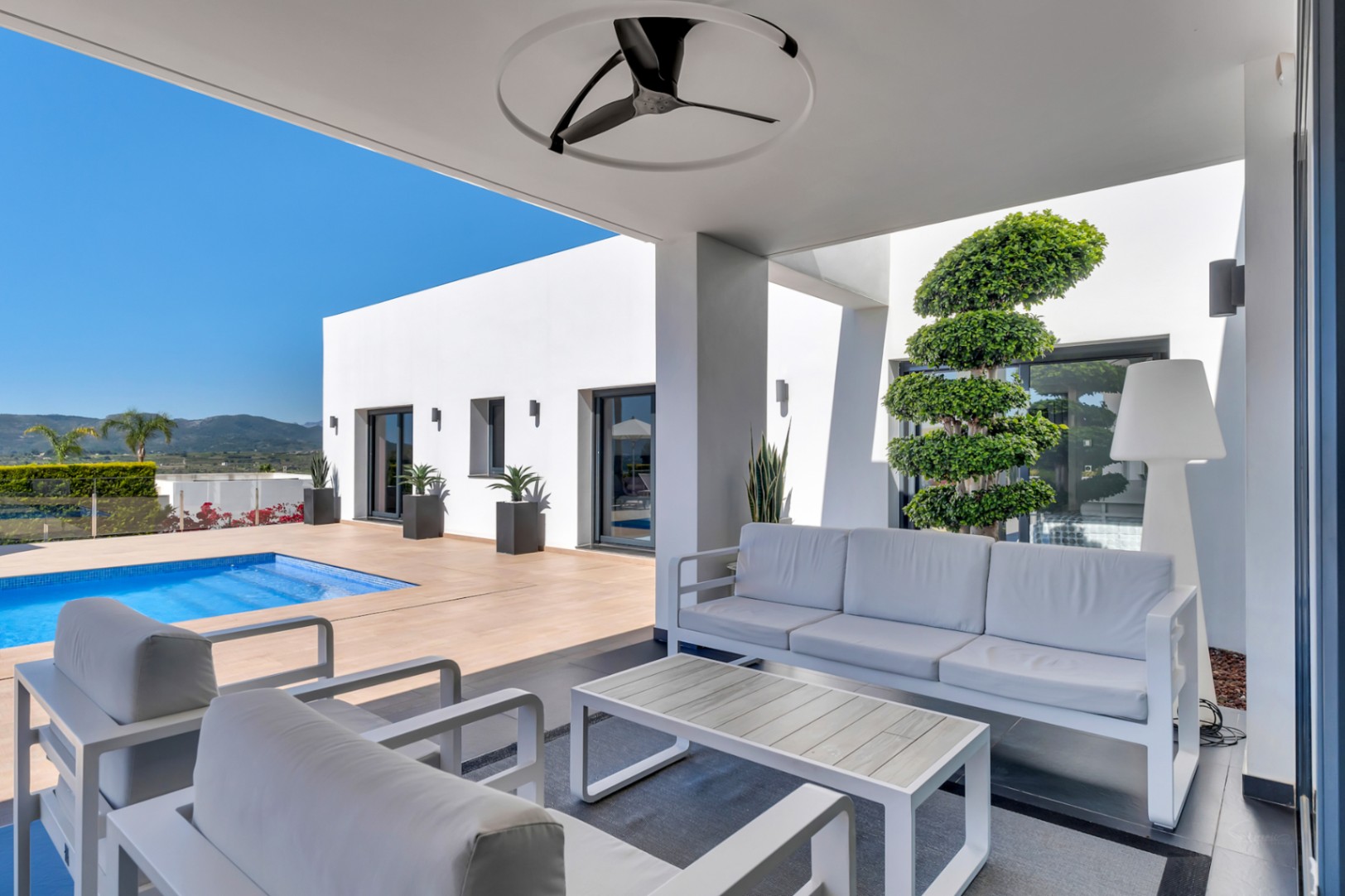 Zona lounge exterior cubierta con mobiliario blanco moderno, ventilador de techo y acceso directo a la piscina. Arquitectura minimalista y vistas a la montaña.