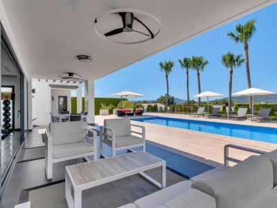 Amplia terraza cubierta con mobiliario lounge blanco, ventiladores de techo y puertas correderas, conectando directamente con la piscina y vistas a las montañas.
