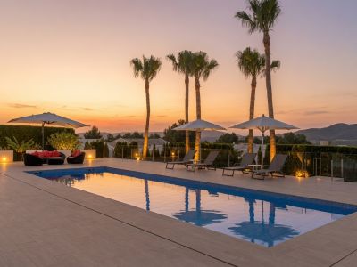 Amplia terraza de piscina exterior con elegantes tumbonas, palmeras iluminadas y zona de estar, enmarcada por un atardecer vibrante y vistas naturales abiertas.