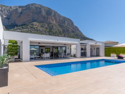 Amplia zona exterior con piscina rectangular de mosaico azul, terraza de baldosas claras y villa moderna blanca con salón y comedor cubiertos integrados, con el Montgó de fondo.