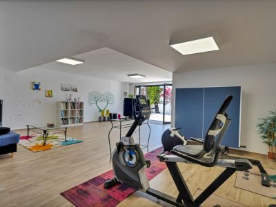 Amplio espacio inferior con equipamiento de gimnasio, una acogedora zona de estar con sofá azul y estantería. Acceso directo al exterior mediante puertas de cristal y suelo de madera clara.