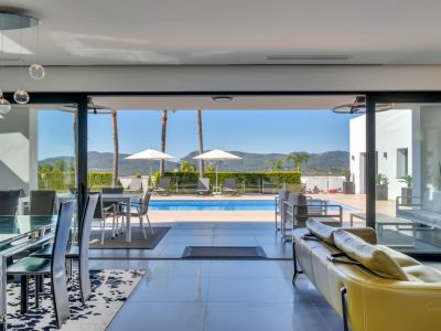 Amplio salón-comedor con mesa de cristal, lámparas colgantes modernas y sofá amarillo, que se abre a la terraza de la piscina con tumbonas y vistas a las montañas lejanas.