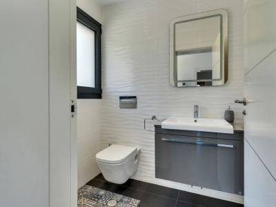 Aseo moderno con inodoro suspendido, mueble de lavabo flotante gris y azulejos blancos texturizados. Destacan el espejo retroiluminado y el detalle de suelo hidráulico.