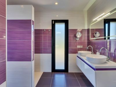 Baño contemporáneo con azulejos morados y blancos, tocador doble flotante con lavabos ovalados y espejo iluminado. Una puerta de cristal esmerilado aporta privacidad.