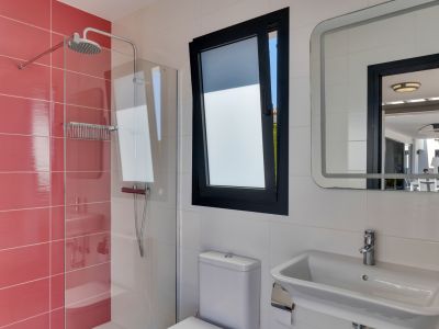 Baño funcional con ducha de plato de lluvia y azulejos rojos, ventana oscilobatiente y espejo iluminado que refleja la terraza exterior.