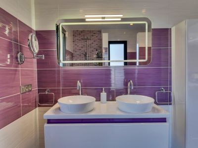 Baño moderno con doble lavabo sobre encimera, azulejos morados horizontales y un espejo iluminado que refleja la ducha con mosaico.