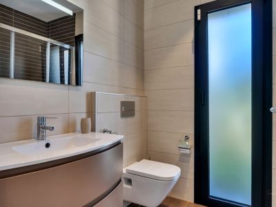 Baño moderno con inodoro suspendido, lavabo con mueble bajo y grifería cromada. Paredes alicatadas en tono neutro y puerta con cristal esmerilado para luz natural.