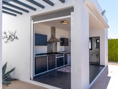 Cocina exterior con gabinetes oscuros y encimeras, equipada con extractor de acero y fregadero, adyacente a un práctico baño exterior y un seto de privacidad.