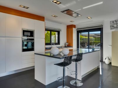 Cocina moderna con gran isla central y asientos, electrodomésticos integrados, armarios blancos y acceso directo al exterior mediante puerta corredera de cristal.
