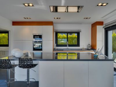 Cocina moderna con mobiliario blanco y encimeras oscuras, destacando una isla central con asientos. Electrodomésticos integrados y luz natural se combinan con vistas a la vegetación exterior.