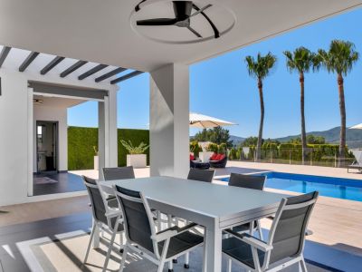 Comedor exterior cubierto con mesa y sillas modernas, ventilador de techo, vistas a la piscina, palmeras y montañas, con un baño exterior adyacente.