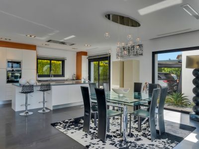 Comedor integrado con cocina moderna de diseño, mesa de cristal y sillas negras, bajo una lámpara colgante contemporánea. Grandes puertas correderas conectan con el exterior.