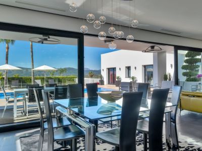 Comedor moderno con mesa de cristal y sillas negras, conectado a una terraza con piscina, tumbonas, palmeras y vistas a la montaña.