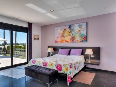 Dormitorio con acceso directo a la terraza a través de grandes puertas correderas de cristal, suelo de baldosas oscuras, edredón floral y una colorida obra de arte abstracta. Vistas al exterior con palmeras.