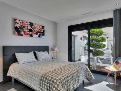 Dormitorio moderno con cama doble y cabecero gris oscuro, acentuado por arte contemporáneo. Puerta corredera de cristal con acceso directo a un patio privado con un árbol escalonado.