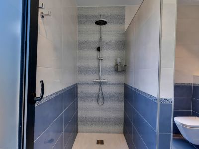 Ducha de obra con azulejos bicolores en azul y blanco, y una franja decorativa de mosaico. Equipada con rociador de lluvia y ducha de mano, separada por mamparas de cristal.