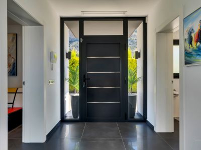 Elegante recibidor con moderna puerta oscura, paneles de cristal laterales y superior, que ofrece luz natural y vistas al exterior y montañas. Suelo de baldosas oscuras e intercomunicador.