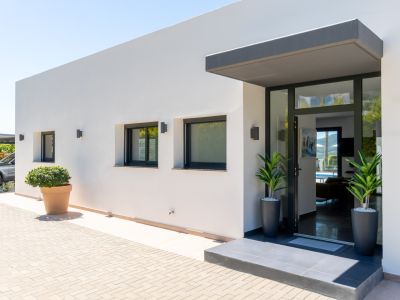 Entrada principal de villa moderna con fachada blanca, puerta acristalada y toldo oscuro. Adoquinado exterior, ventanas con persianas y aparcamiento cubierto visible.