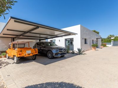 Exterior de villa moderna con doble aparcamiento cubierto de estructura oscura, albergando un SUV Mercedes y un coche descapotable naranja, sobre un camino pavimentado.