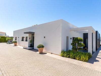 Exterior de villa moderna con fachada blanca, entrada pavimentada y paisajismo minimalista. Destacan un árbol esculpido, pérgola y vistas a las colinas.