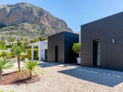 Exterior moderno de la villa con fachadas de azulejos oscuros y detalles en blanco, enmarcada por el imponente Montgó. Jardín de grava con plantas mediterráneas.