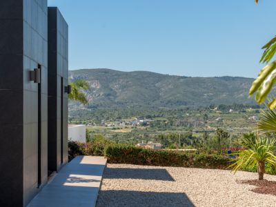 Fachada exterior con revestimiento de paneles oscuros y apliques de pared, sendero pavimentado, jardín de grava y amplias vistas a las montañas del Montgó.