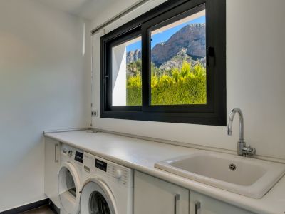 Lavadero funcional con encimera blanca, fregadero integrado y lavadoras, destacando una ventana con vistas a setos verdes y la majestuosa montaña del Montgó.